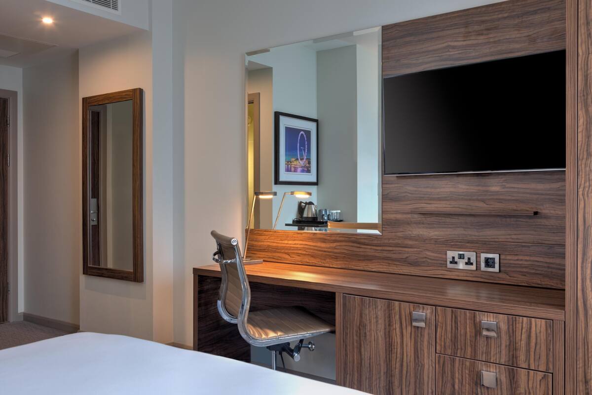 A hotel room with a bed, a wooden desk, a chair, a mirror, a television, and a framed picture on the wall.