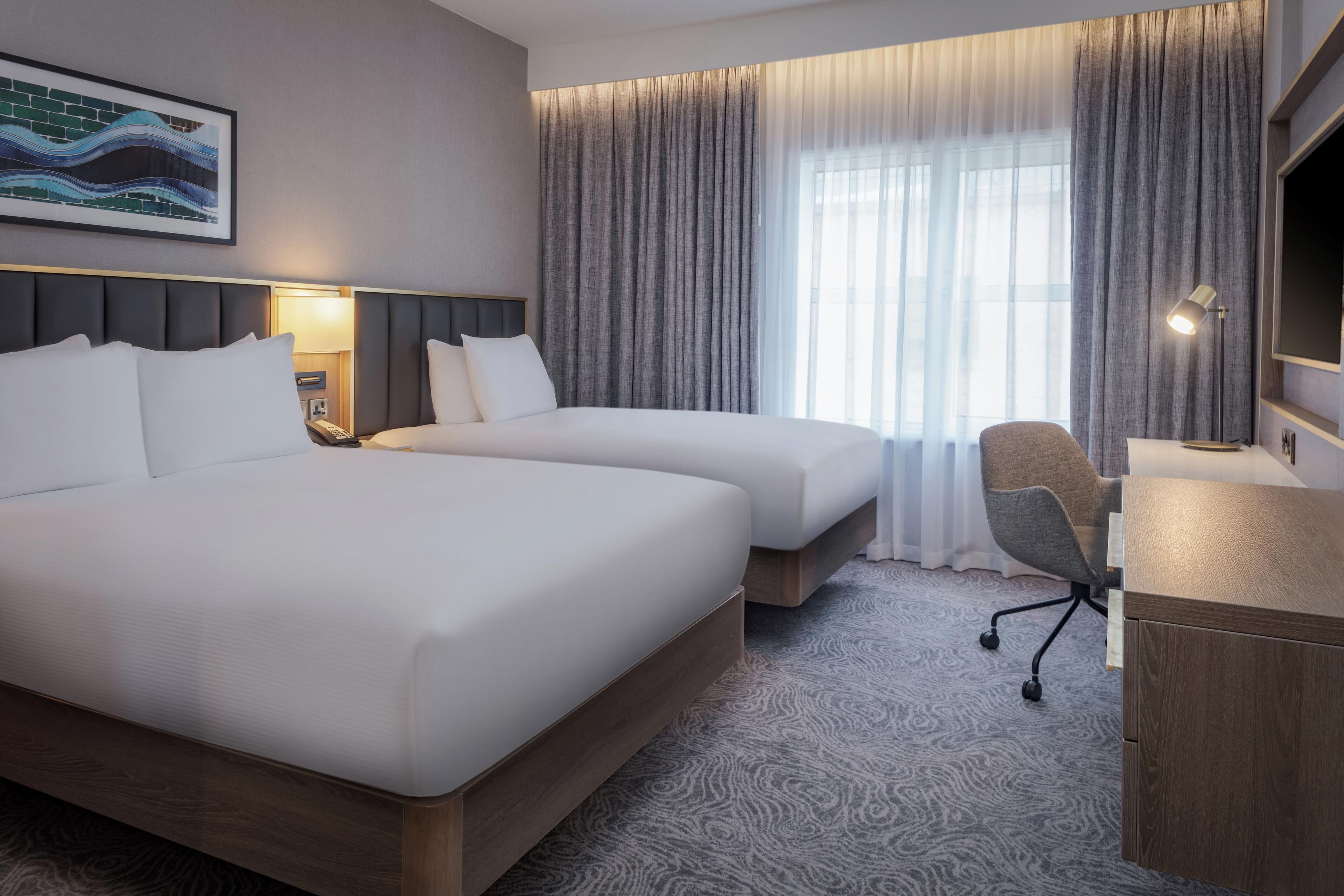 Une chambre d’hôtel avec deux lits, un bureau, une chaise, une lampe et une grande fenêtre avec des rideaux.
