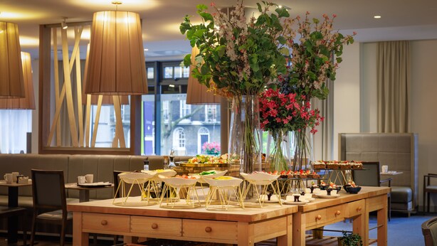 A dining area with a wooden table set for a buffet, surrounded by chairs and a vase of flowers. - DoubleTree by Hilton London Angel Kings Cross (London)