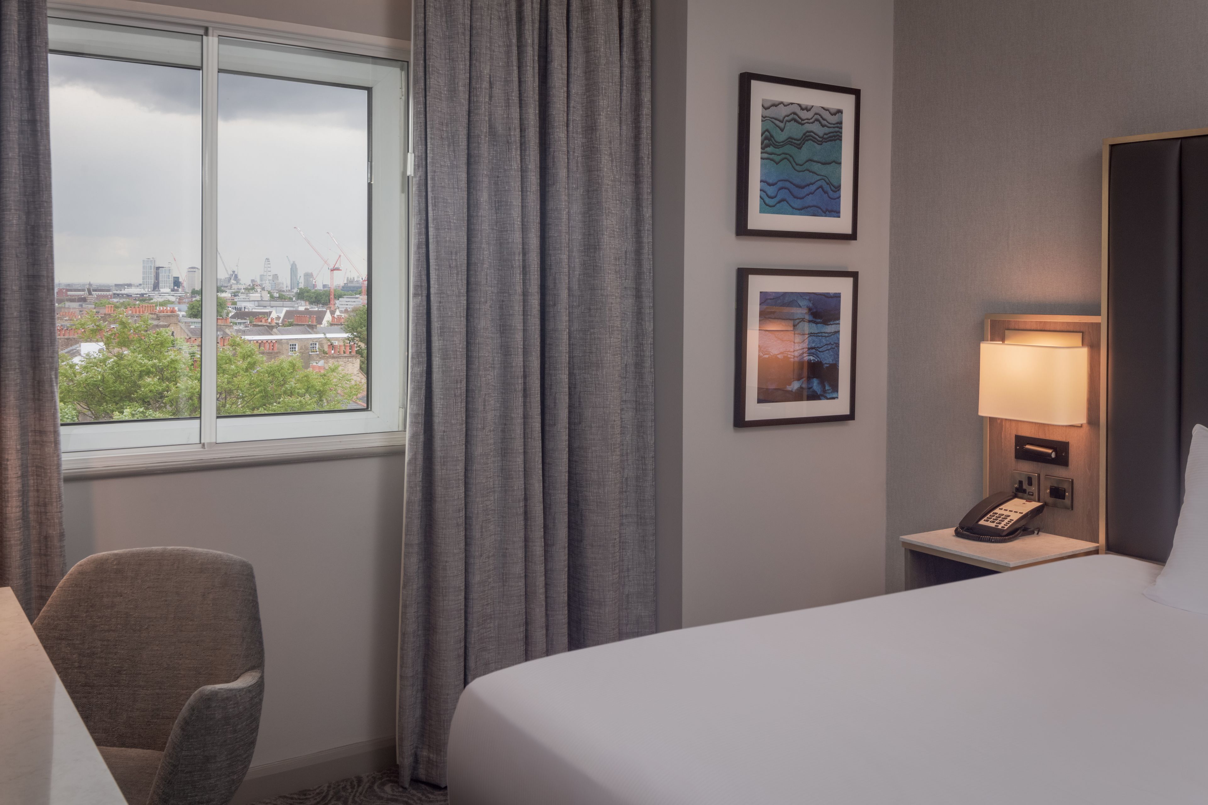 Ein Hotelzimmer mit Bett, Stuhl, Schreibtisch mit Lampe und zwei gerahmten Bildern an der Wand.