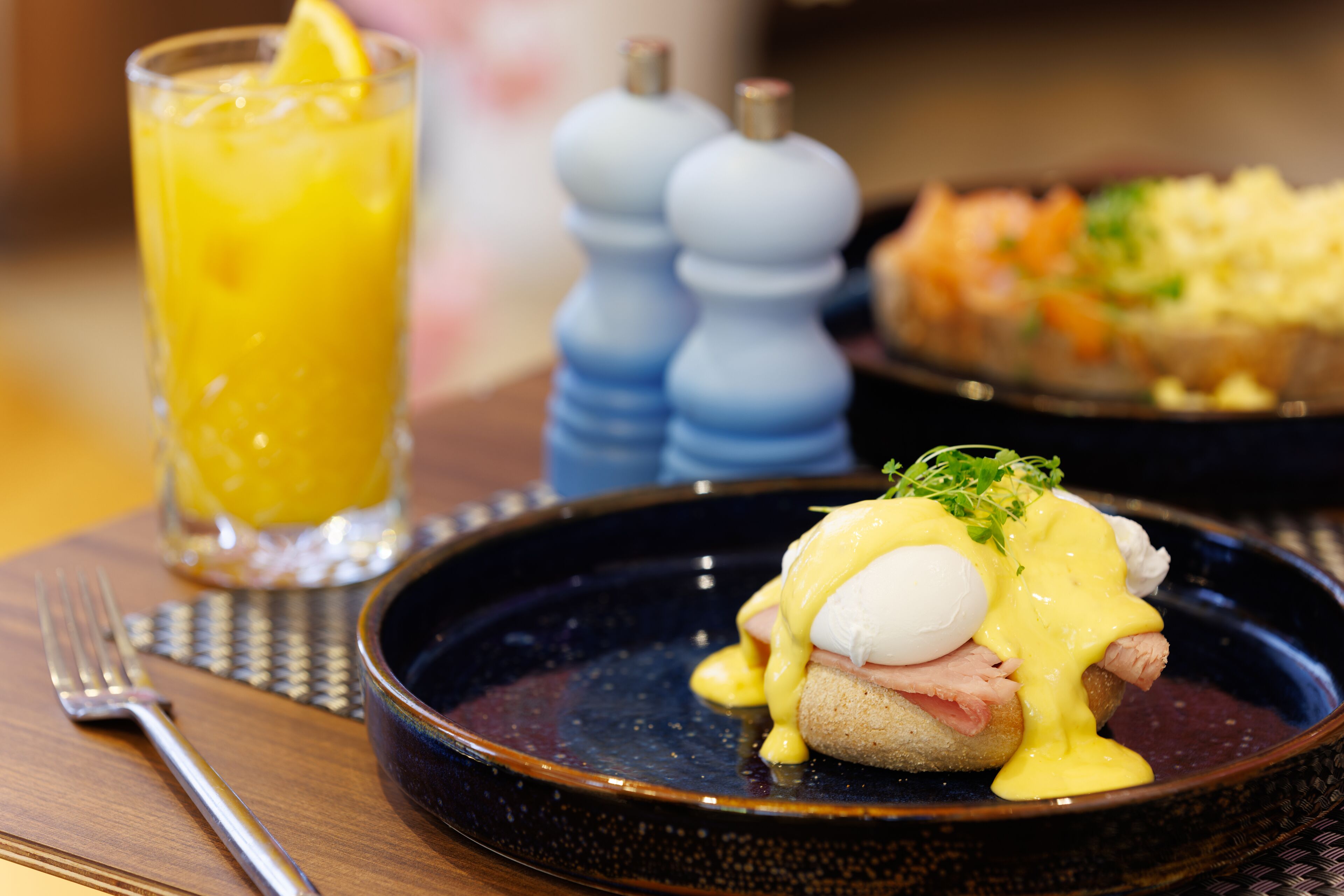 A dining table with a plate of eggs benedict, a glass of orange juice, and a salt and pepper shaker.