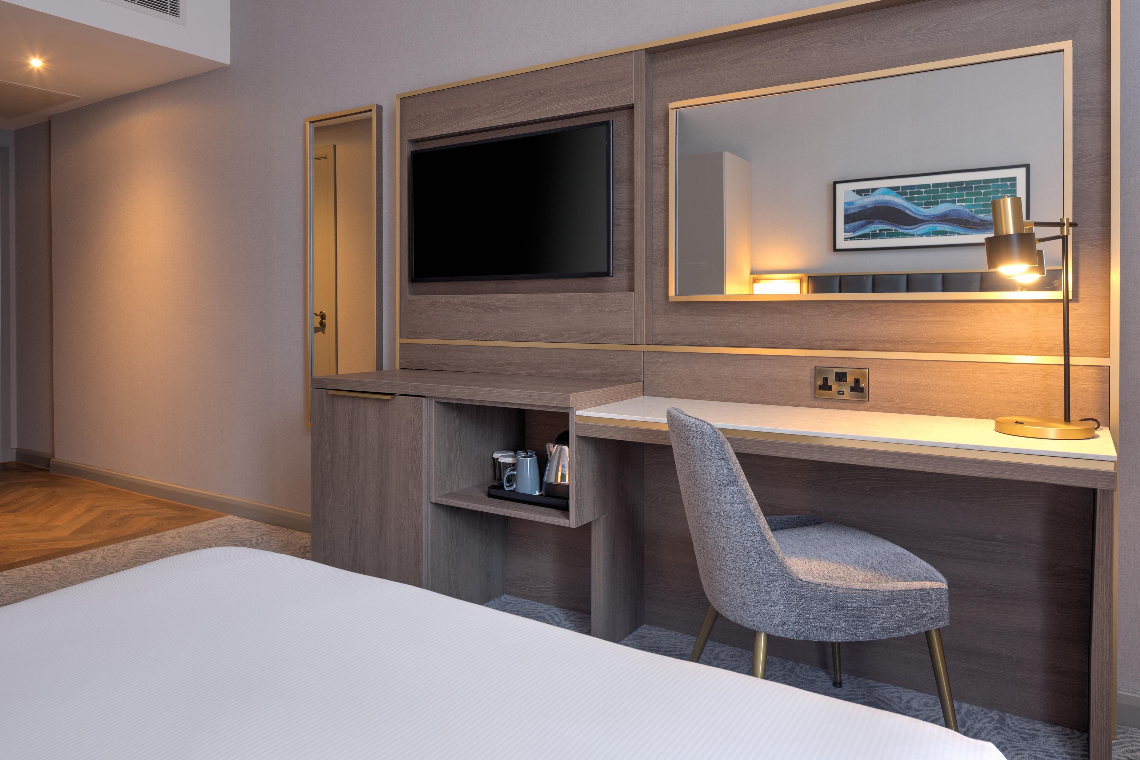A modern hotel room with a bed, a desk with a chair, a television, and a mirror.