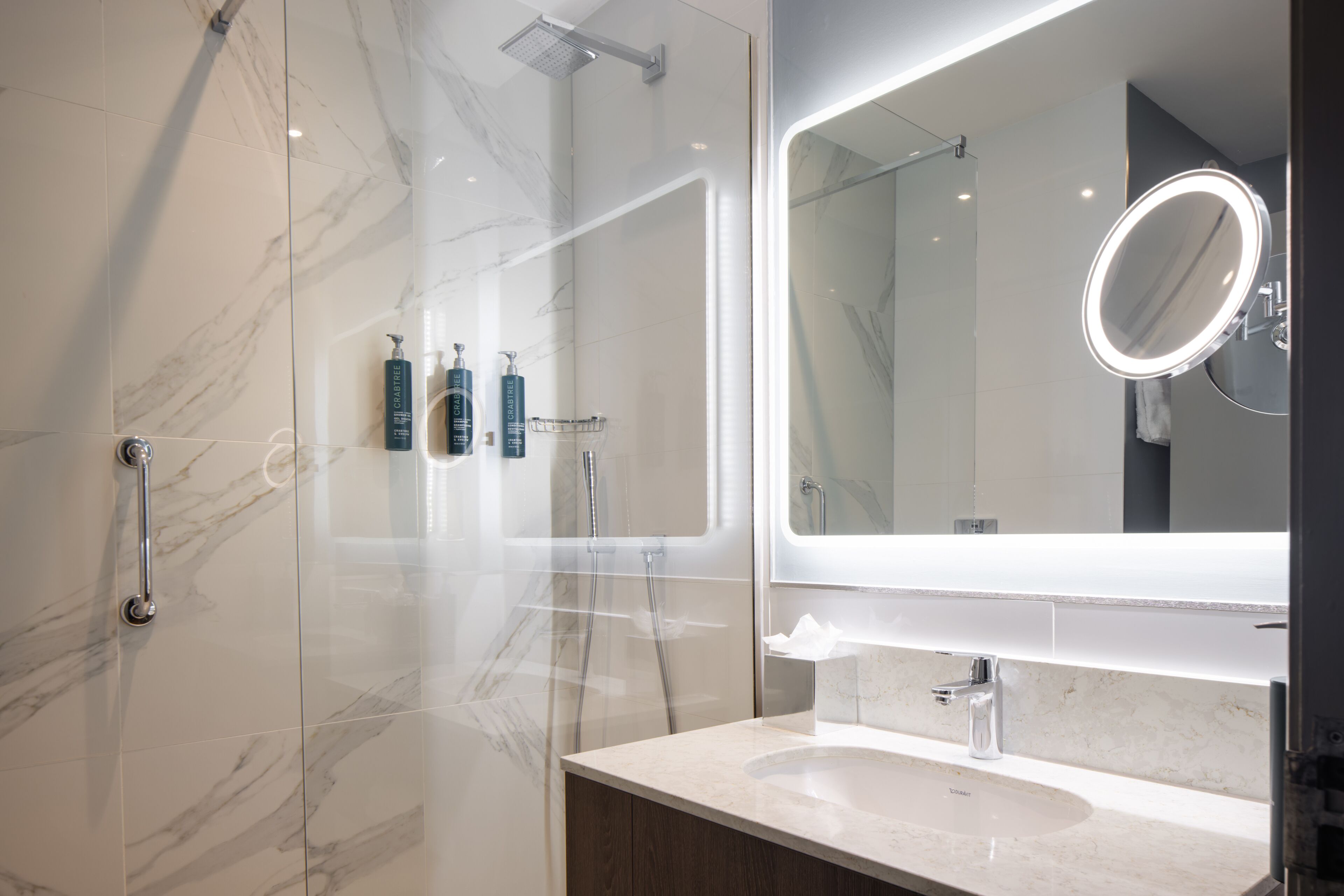 A modern bathroom with a glass-enclosed shower, a large mirror with a round frame, and a marble-tiled vanity.