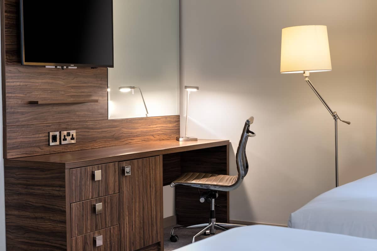 A hotel room with a wooden desk, a chair, a TV mounted on the wall, a lamp, and a bed.