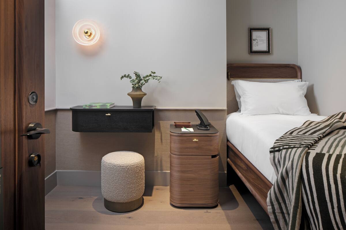 Un cuarto de hotel ordenado con cama, mesita de noche y un pequeño taburete.