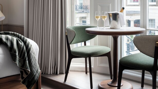 A room with a view of a cityscape through large windows, a round table with two chairs, and a bottle of champagne. - Montcalm Mayfair Townhouse (London)