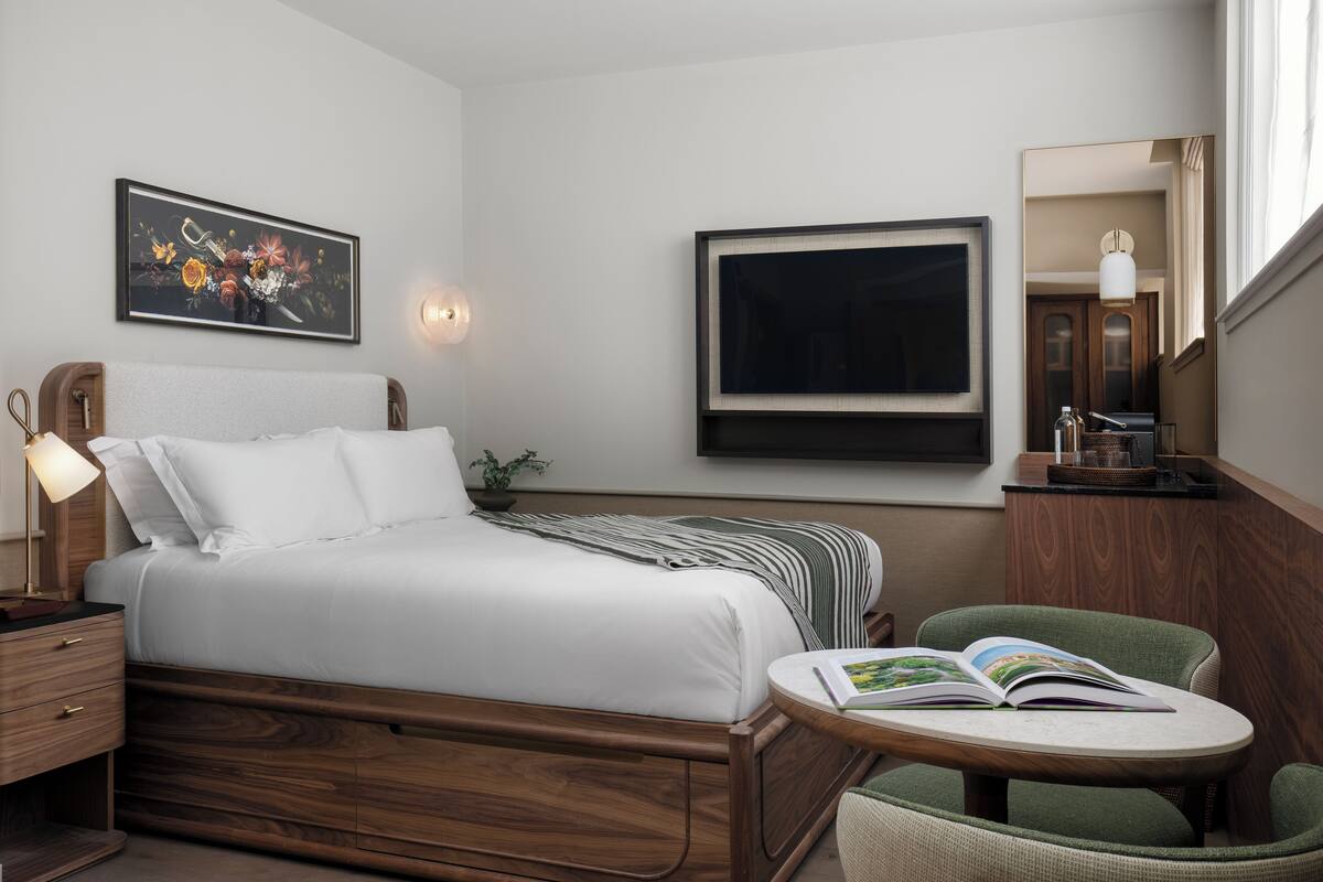 Una habitación de hotel con una cama de madera, una mesita de noche con una lámpara, una silla verde, una mesa redonda con un libro y un cuadro en la pared.