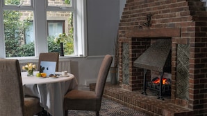 Una habitación con chimenea de ladrillo, una mesa de comedor con sillas, un florero con flores y vistas a través de una ventana.