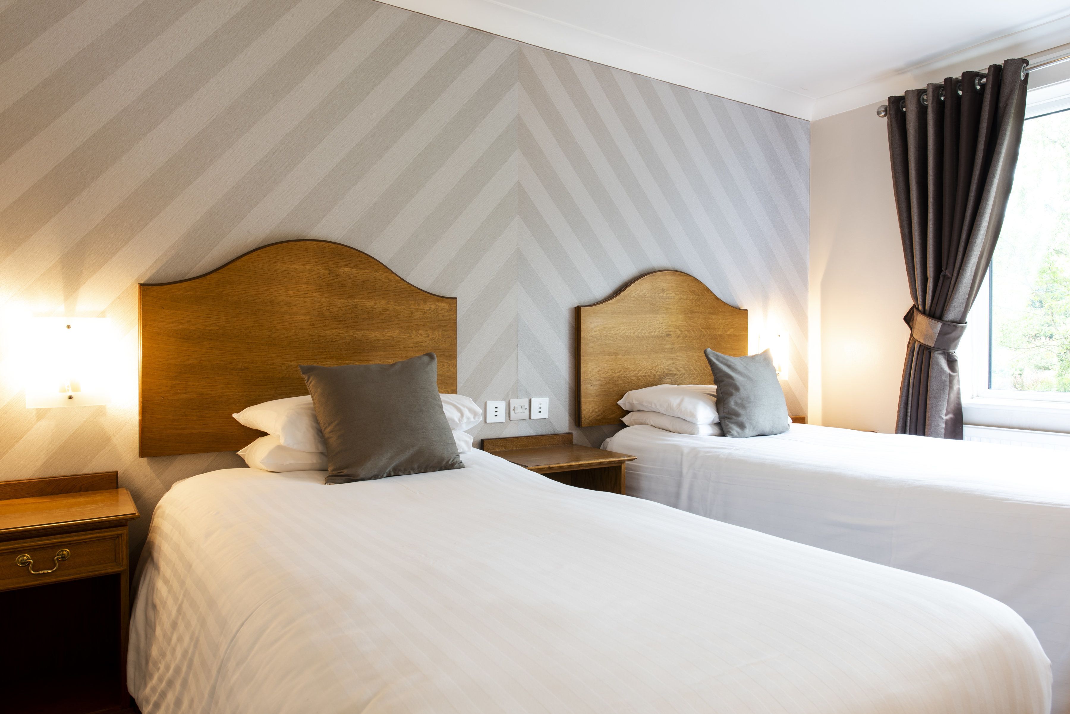 A hotel room with two beds, wooden headboards, white bedding, and a window with curtains.