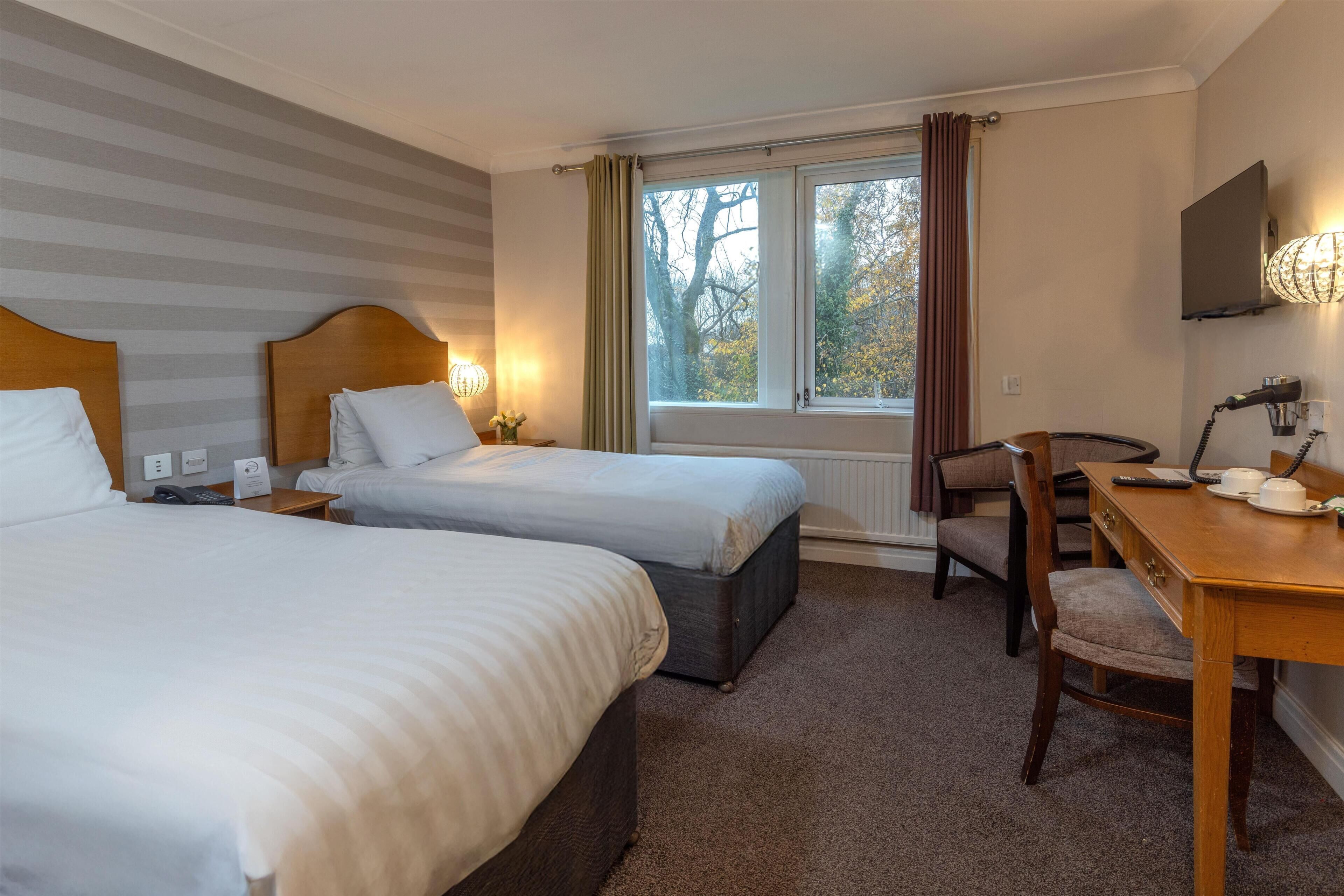 A hotel room with two beds, a desk, a chair, a TV, and a window with curtains.