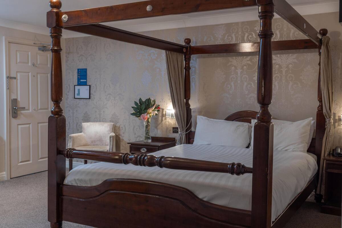A four-poster bed with a wooden frame, a bedside table with a lamp, a chair, and a wall-mounted picture.