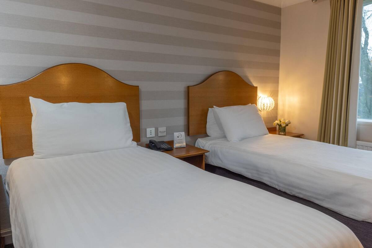 A hotel room with two single beds, wooden headboards, a bedside table with a lamp, a phone, and a sign.