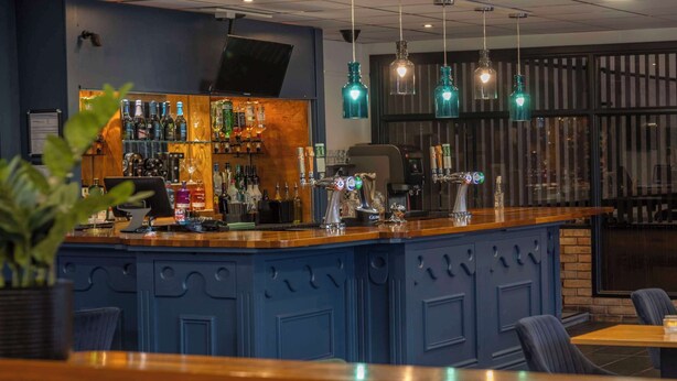 Un espace bar avec un comptoir bleu, des étagères remplies de bouteilles d’alcool et des lampes suspendues vertes.