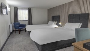 Une chambre d’hôtel avec deux lits, un bureau, une chaise, un radiateur et une fenêtre avec des rideaux.