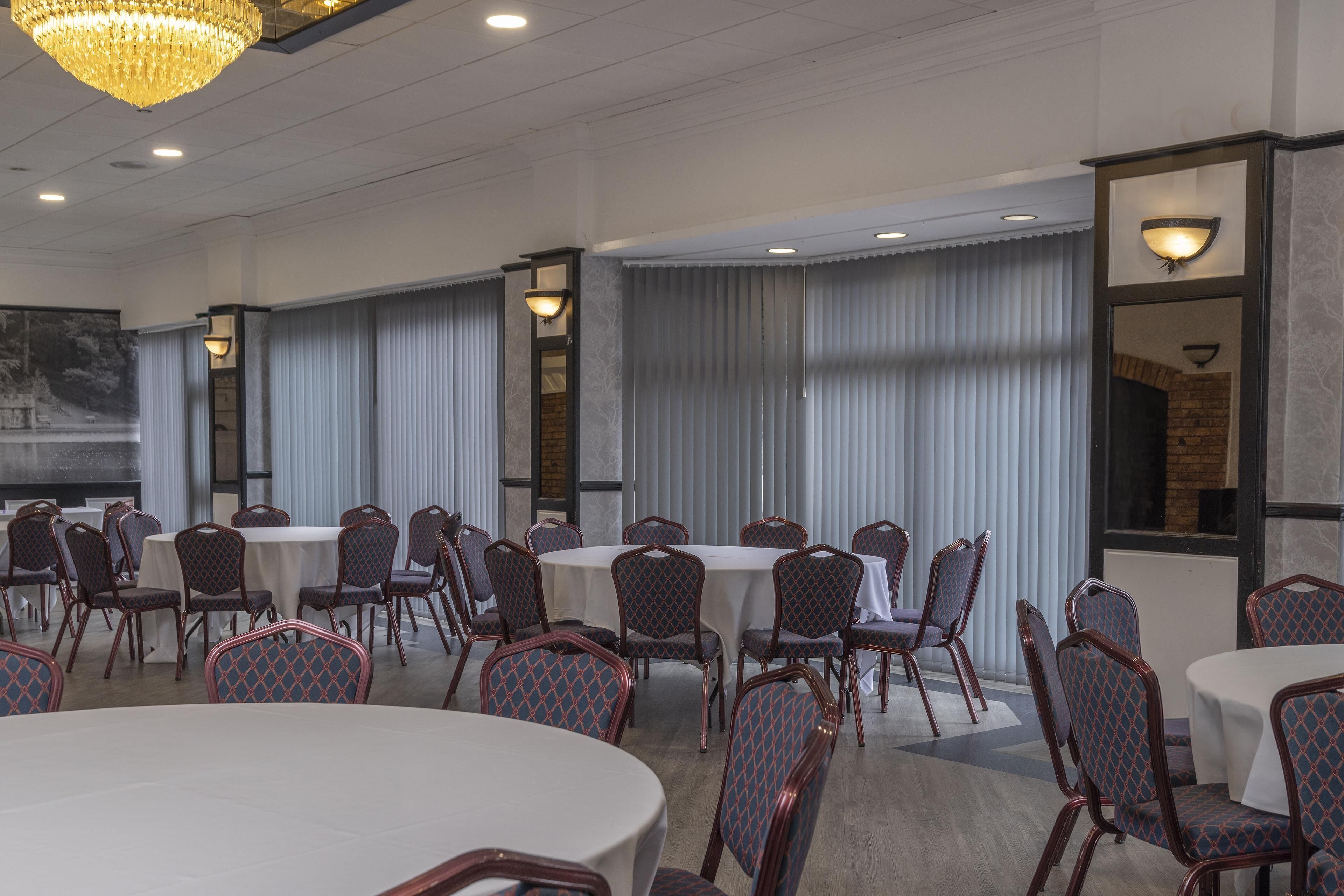 A conference room with round tables and chairs, large windows with vertical blinds, and a chandelier.