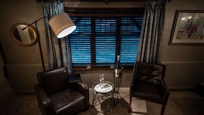 Une pièce avec deux fauteuils en cuir, une petite table ronde sur laquelle se trouve un verre à vin, un lampadaire et une fenêtre ornée de rideaux à motifs.