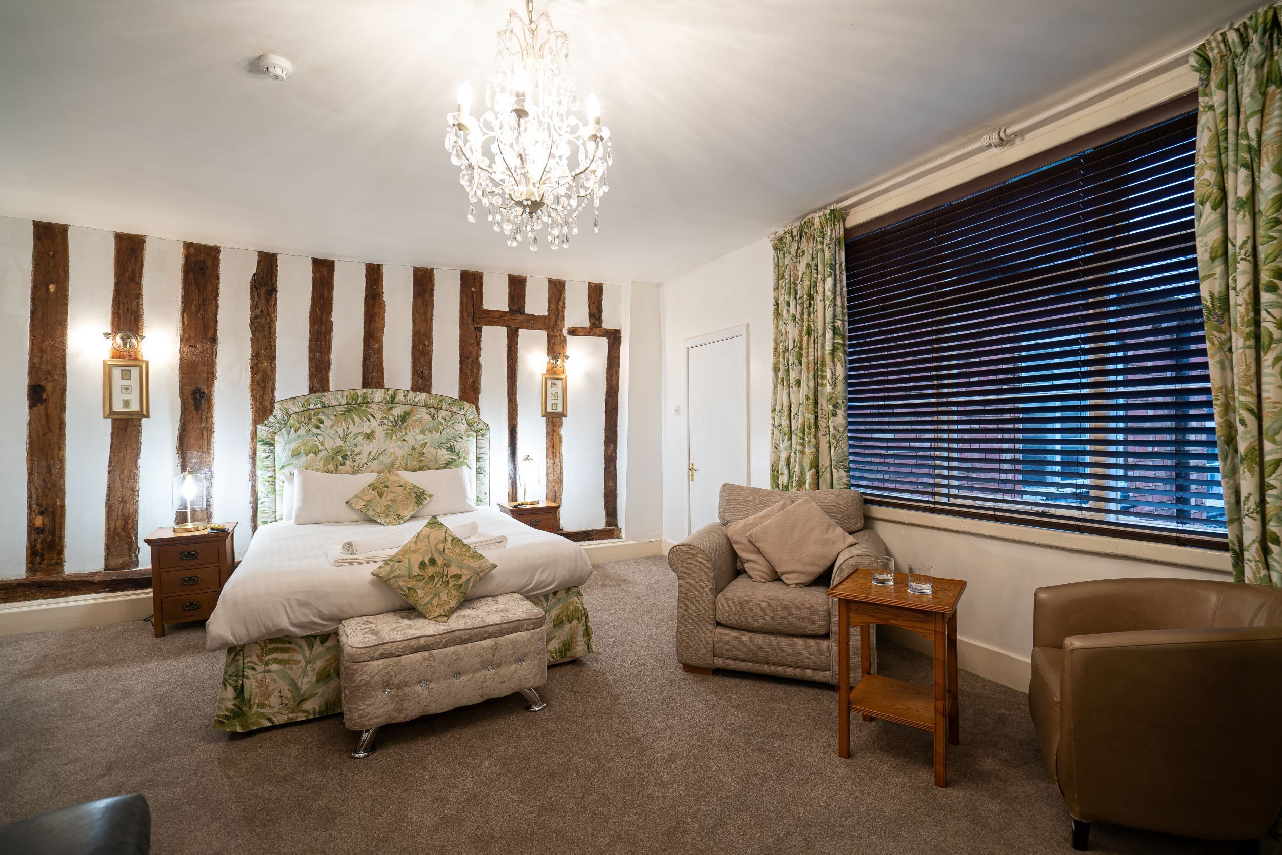 A bedroom with a large bed, a chandelier, a wooden wall panel, a window with blinds, and a nightstand with a lamp.