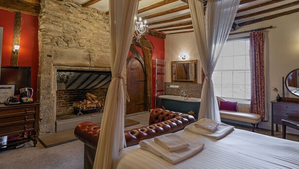 Une chambre avec une cheminée en pierre, un grand lit à baldaquin, un banc en bois et une salle de bain avec une baignoire et un miroir.