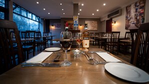 Une table de restaurant, dressée avec des verres à vin, une bougie et une bouteille de spiritueux, entourée de chaises et de tables en bois.