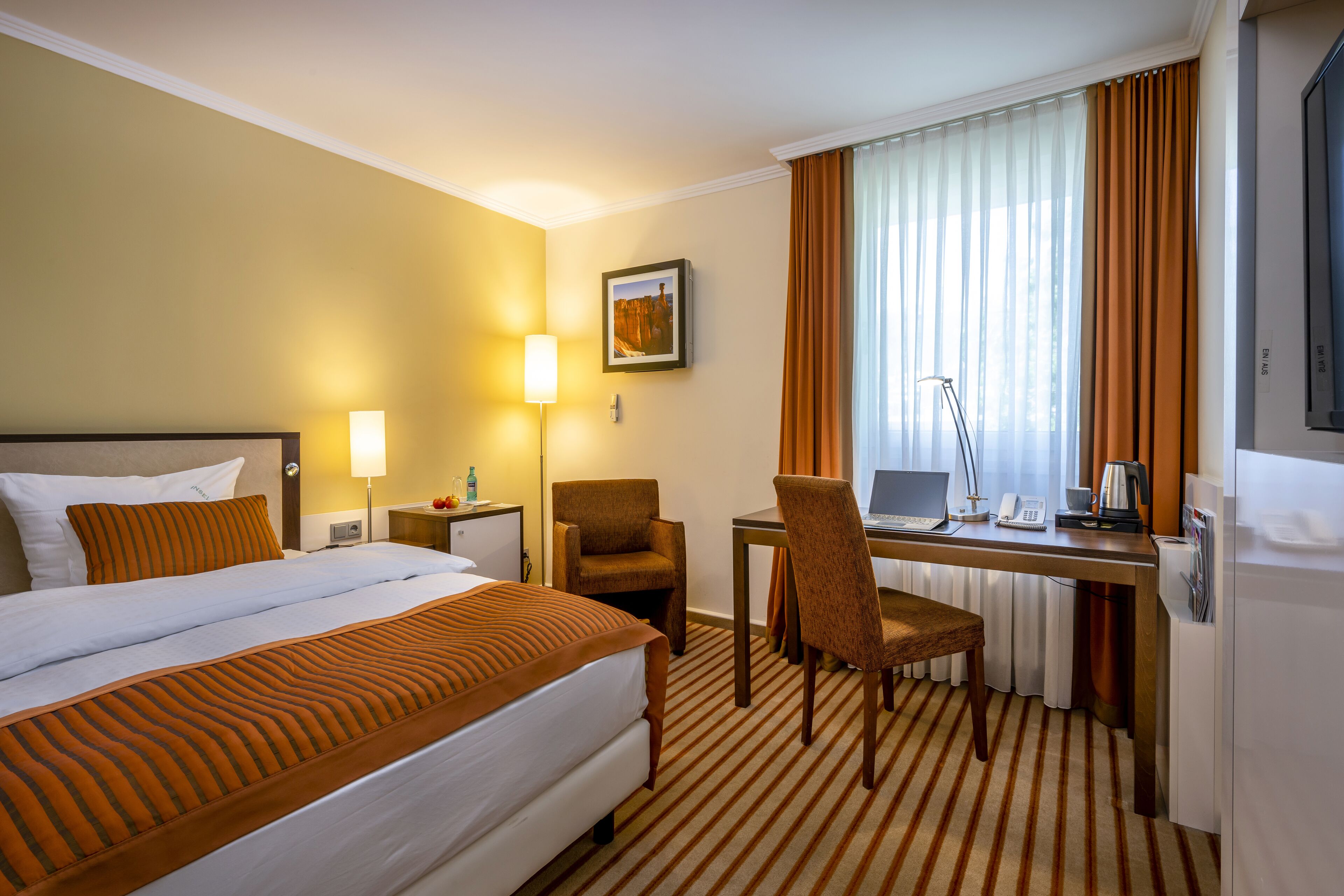 Ein Hotelzimmer mit einem Bett, einem Schreibtisch, einem Stuhl, einer Lampe, einem Laptop und einem Fenster mit Vorhängen.
