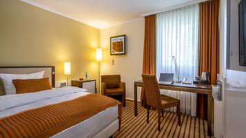 Ein Hotelzimmer mit einem Bett, einem Schreibtisch, einem Stuhl, einer Lampe, einem Laptop und einem Fenster mit Vorhängen.