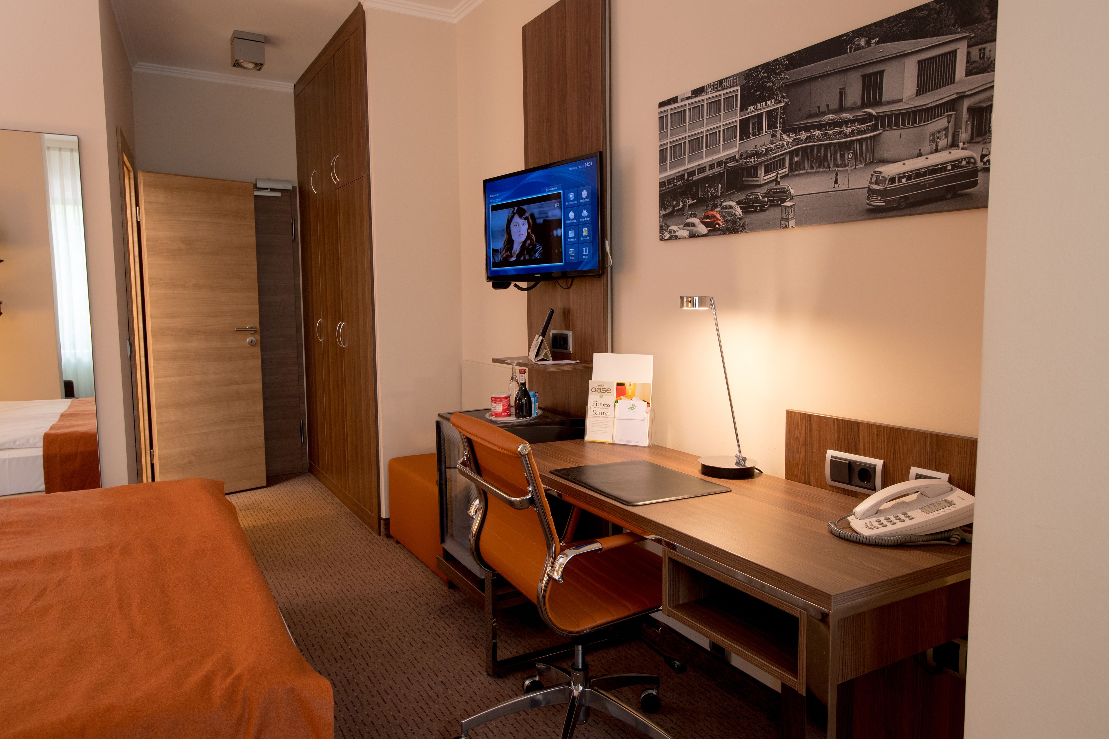 Ein Hotelzimmer mit einem Bett, einem Schreibtisch mit Computer, einem Sessel, einem Telefon und einem an der Wand befestigten Fernseher.