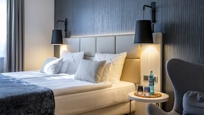 Een moderne hotelkamer met een groot bed, twee bedlampen, een klein tafeltje met glazen en een fles, en een grijze fauteuil.