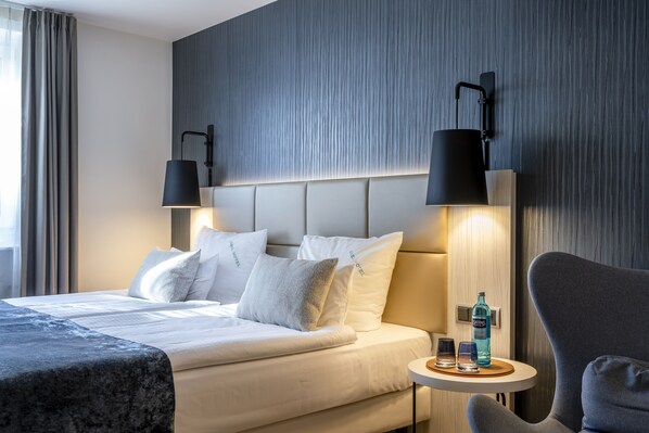 A modern hotel room with a large bed, two bedside lamps, a small table with glasses and a bottle, and a grey armchair.