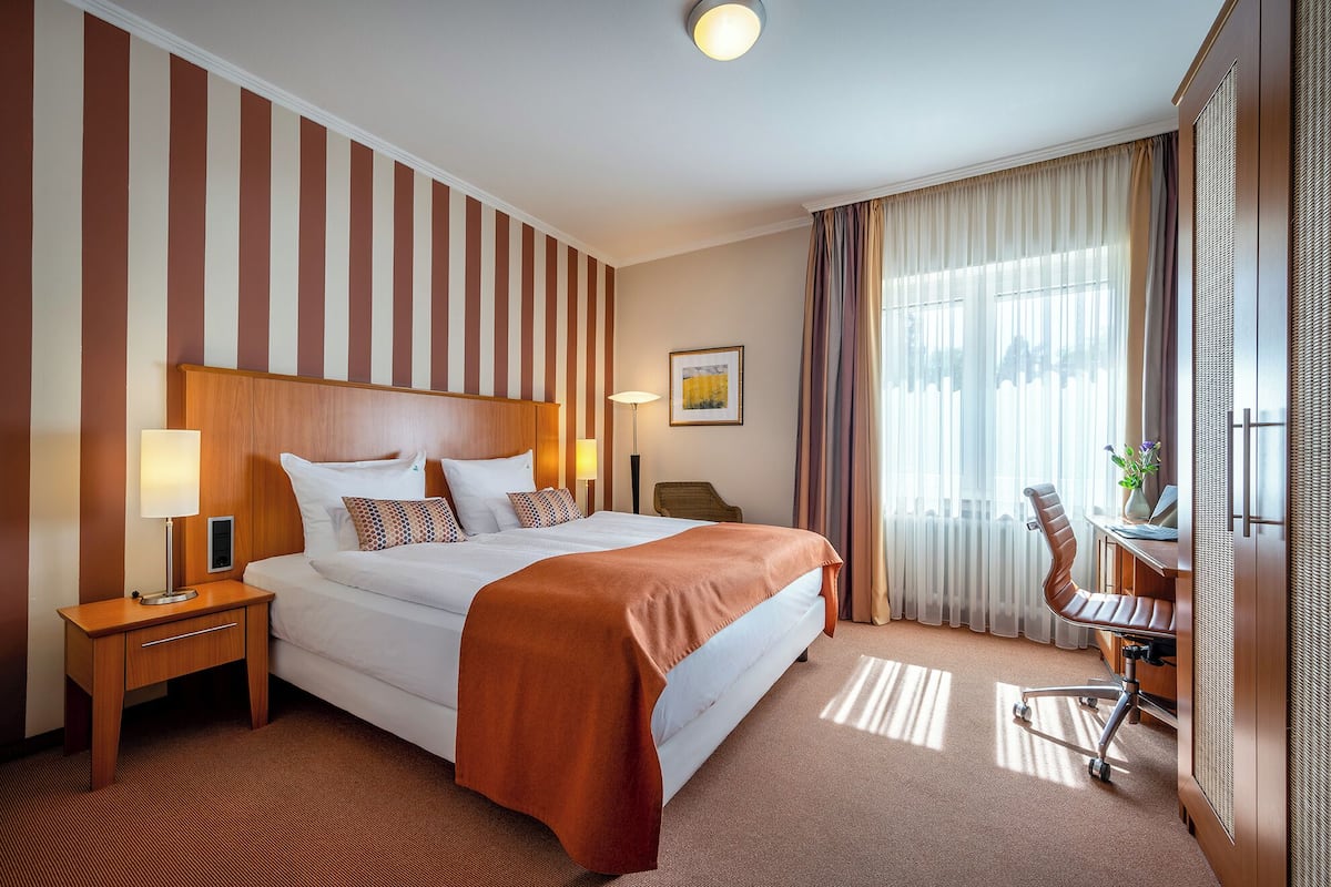 Une chambre d’hôtel avec un grand lit, un bureau avec une chaise, une lampe et une fenêtre avec des rideaux.