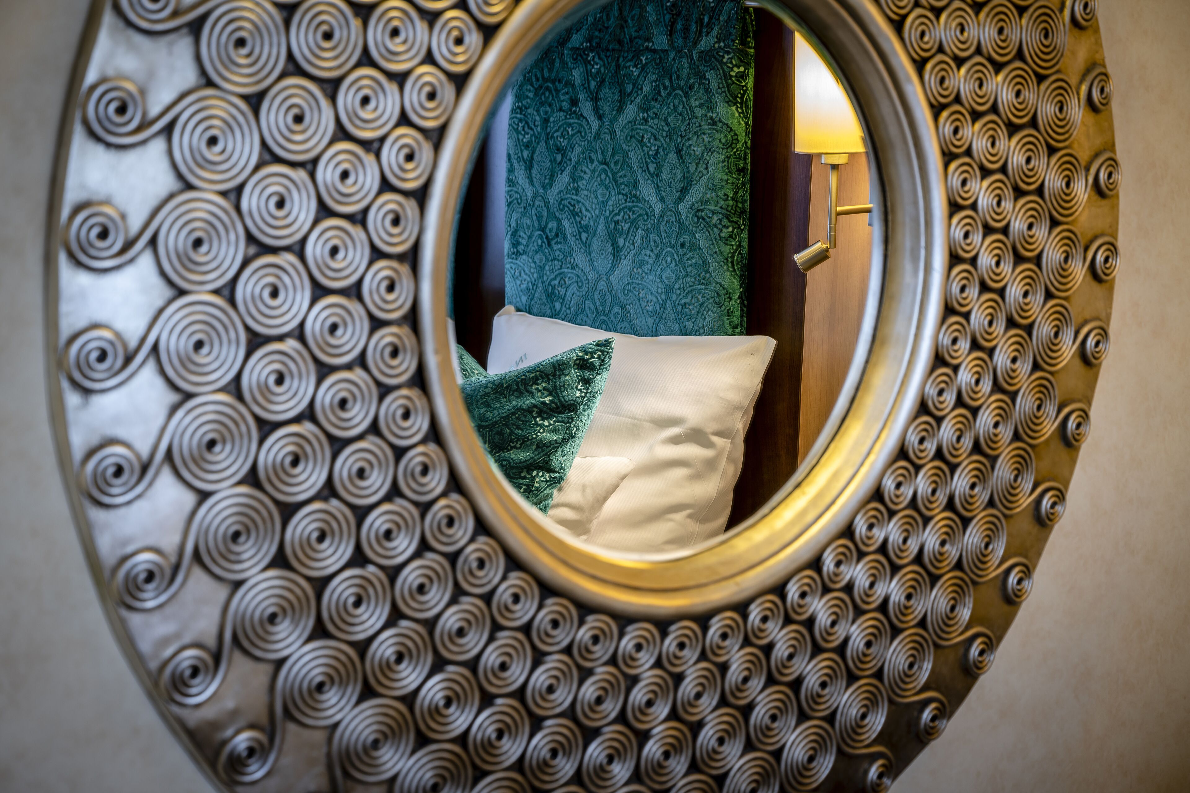 A circular mirror with a decorative spiral pattern, reflecting a room with a green textured wall and a white pillow.