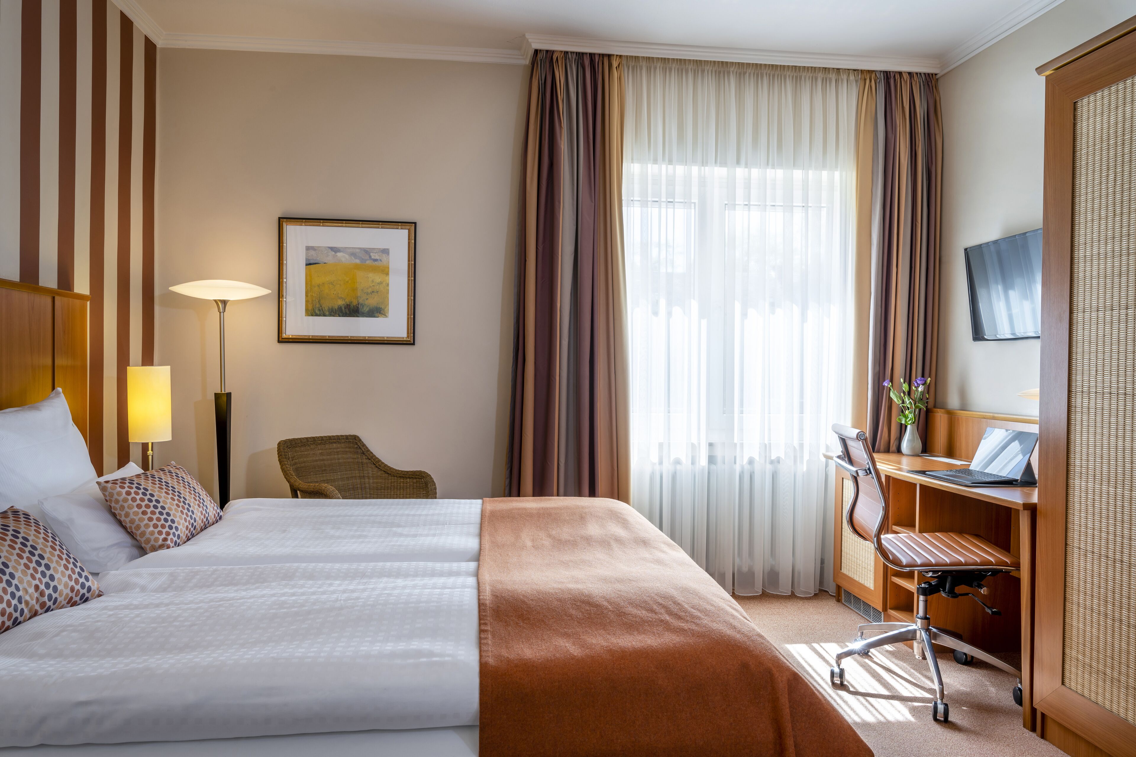 A hotel room with a large bed, a desk with a laptop, a chair, and a painting on the wall.