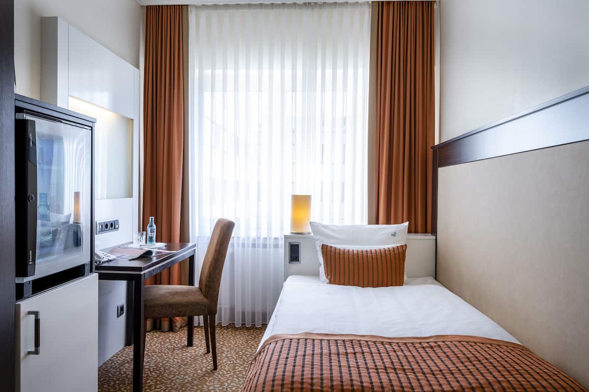 Une chambre d’hôtel comprenant un lit, un bureau avec une télévision, une chaise, une table de chevet avec une lampe et une fenêtre avec des rideaux.