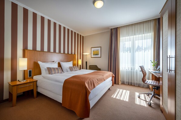 Habitación de hotel con una cama grande, un escritorio con silla, una lámpara y una ventana con cortinas.