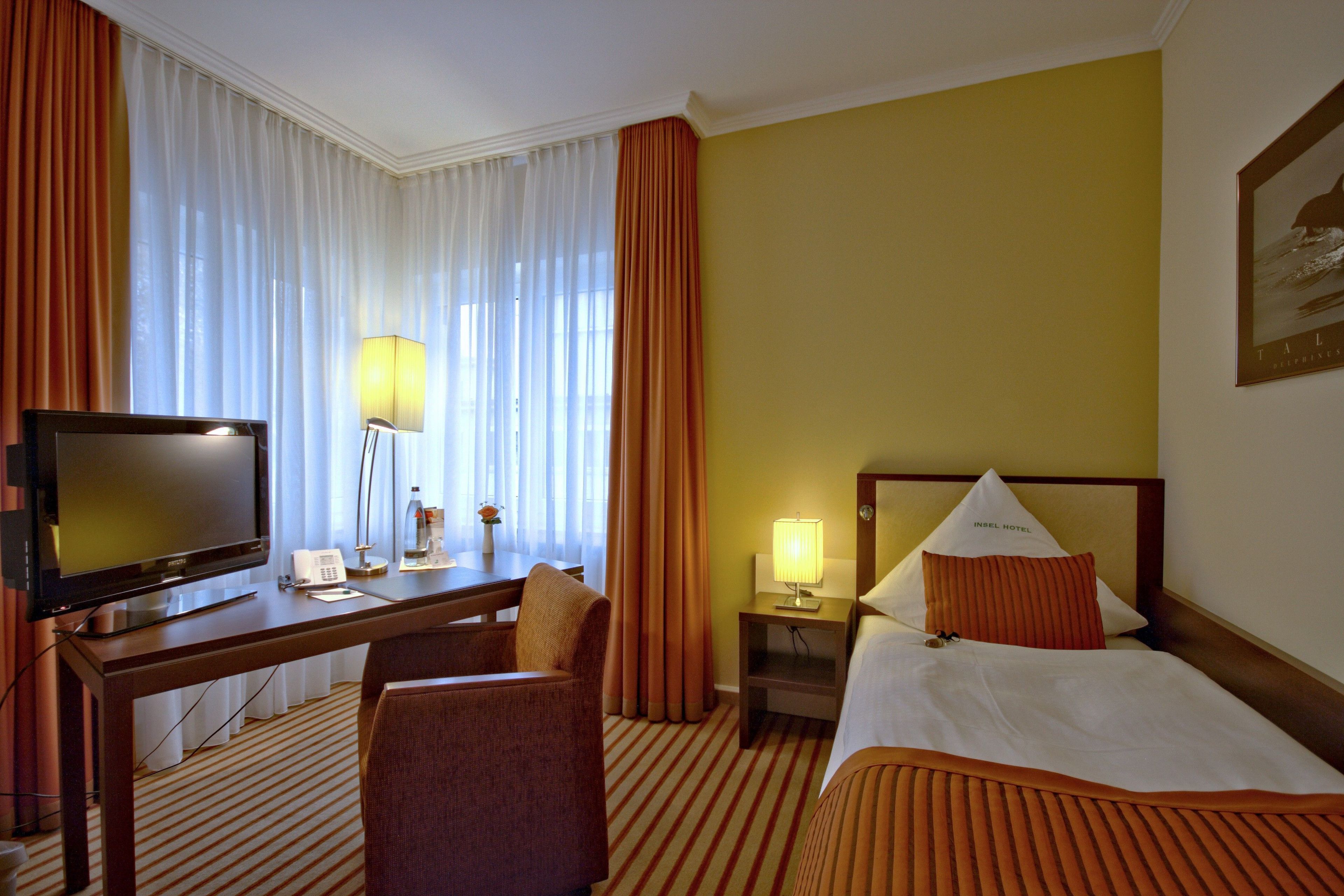 A hotel room with a bed, a desk with a television, a chair, a nightstand with a lamp, and a window with curtains.