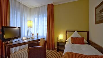 A hotel room with a bed, a desk with a television, a chair, a nightstand with a lamp, and a window with curtains.
