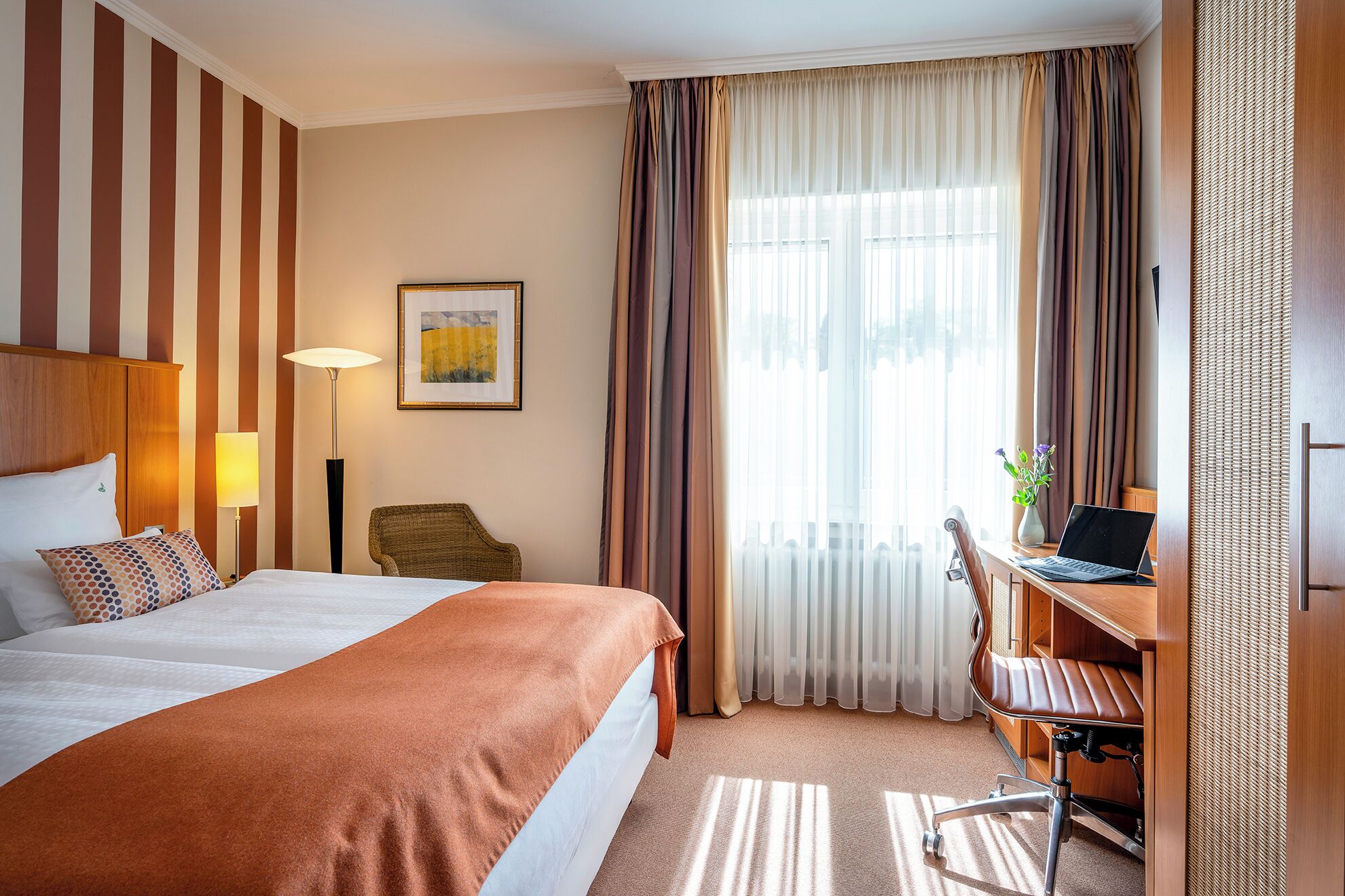 A hotel room with a large bed, a desk with a laptop, a chair, a lamp, and a framed picture on the wall.