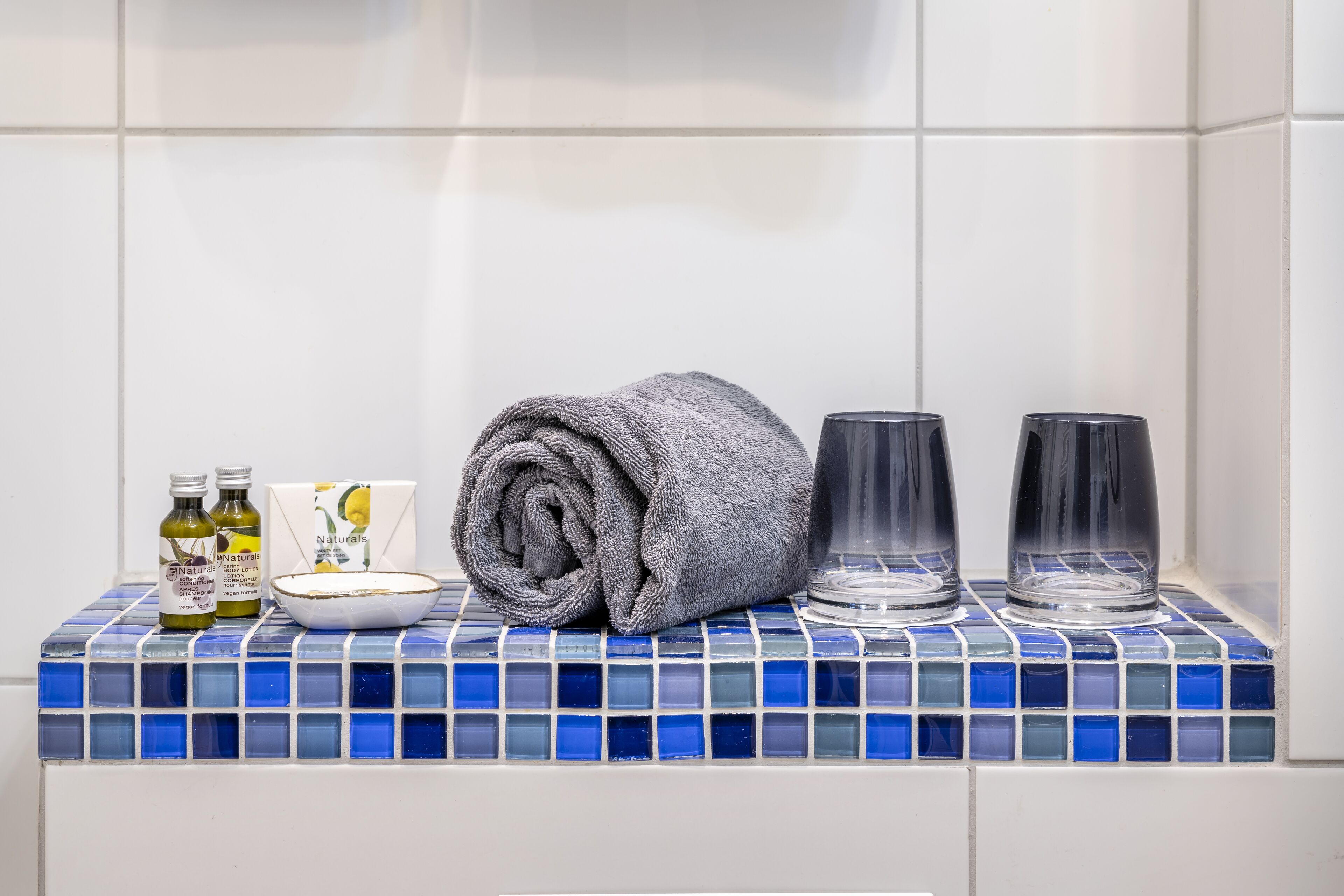 A bathroom with a tiled sink, two bottles of lotion, a rolled towel, and two glass containers.