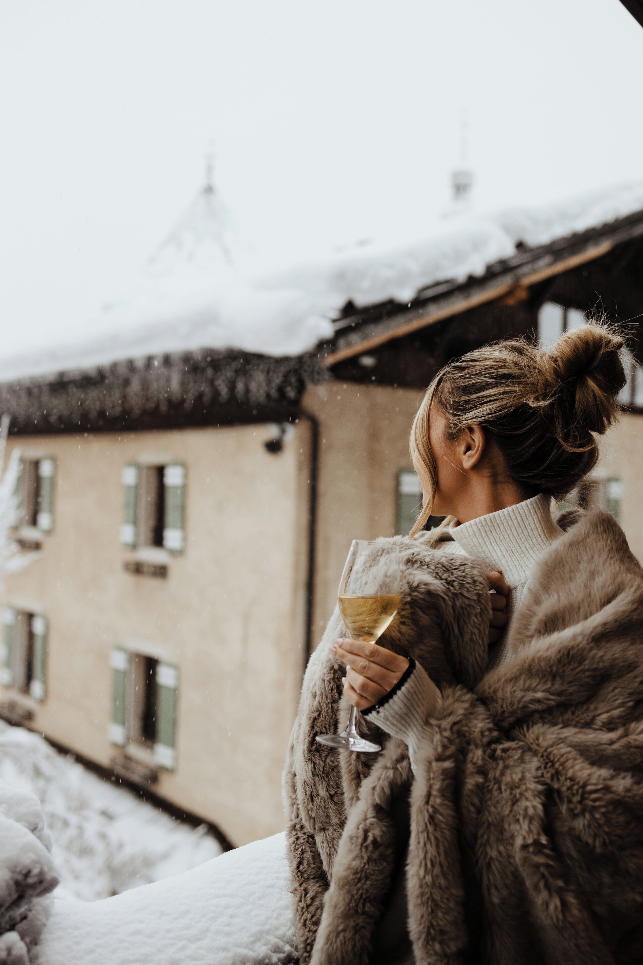 A person in a fur coat holding a glass of wine, standing in front of a building with snow on the roof and windows.