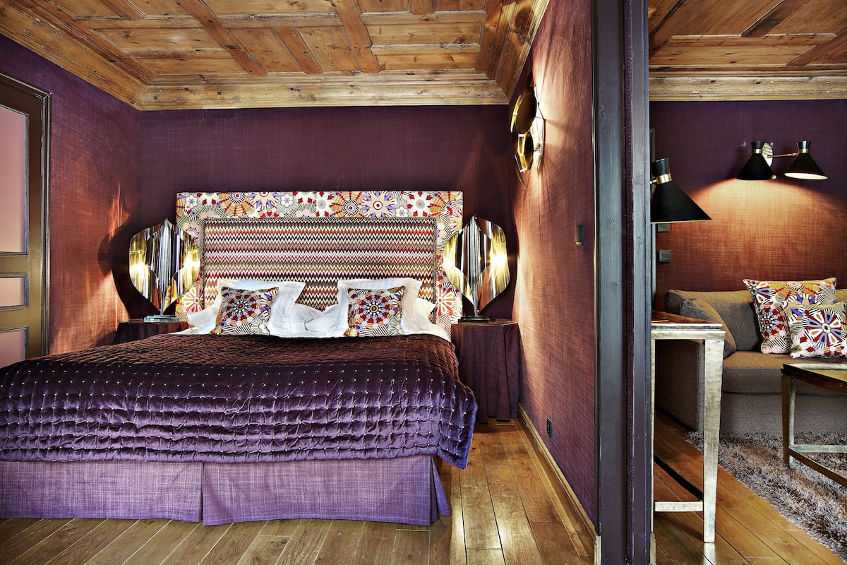 A bedroom with a wooden ceiling, a bed with a purple bedspread, two bedside lamps, and a patterned headboard.