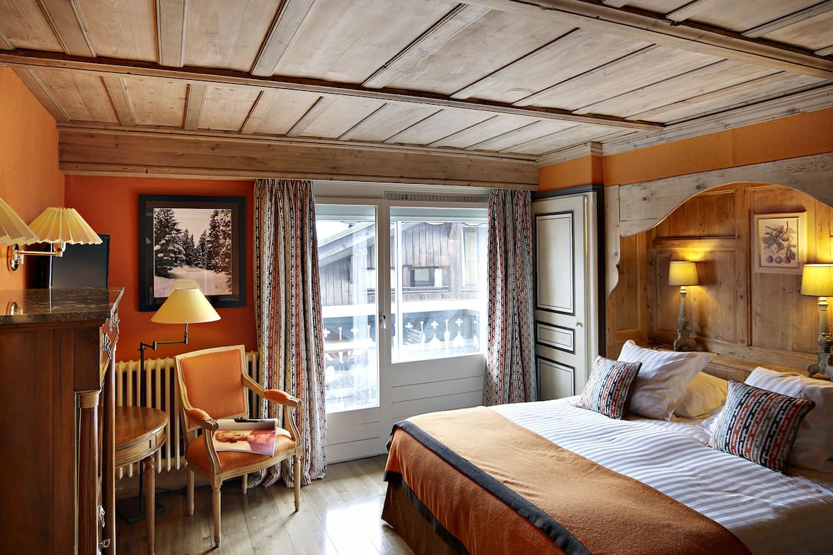 A hotel room with a wooden ceiling, a bed with striped bedding, a chair, a desk with a lamp, and a window with curtains.
