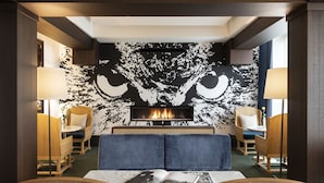 Una habitación de hotel moderna con un gran mural en la pared de la cara de un búho, una chimenea y un área de descanso con sofás tapizados en azul y sillas de madera.