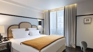 Habitación de hotel con una cama grande, una mesita de noche con lámpara, una silla y una ventana con cortinas.