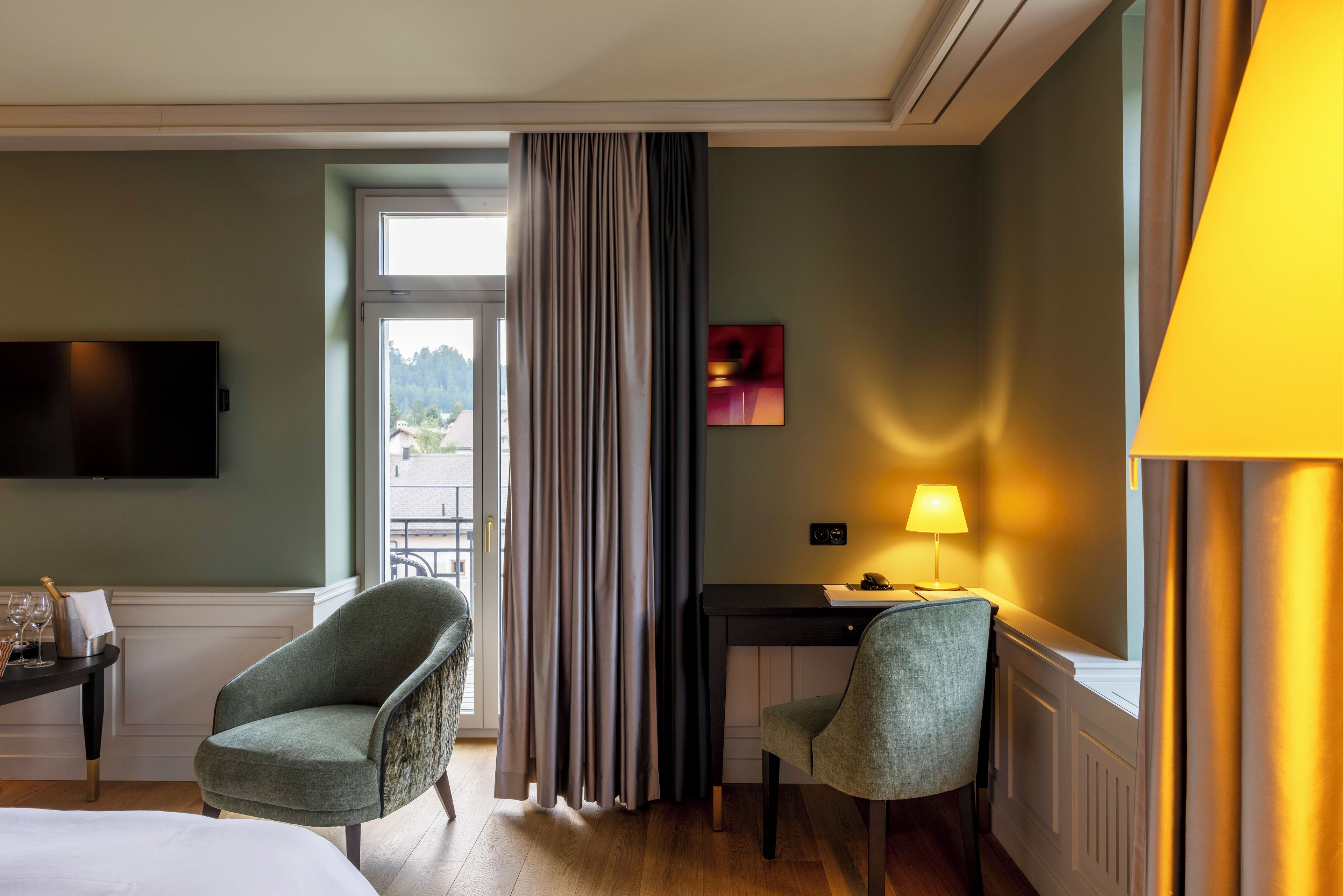 A hotel room with a bed, two armchairs, a desk with a lamp, a TV, and a view of the outdoors.