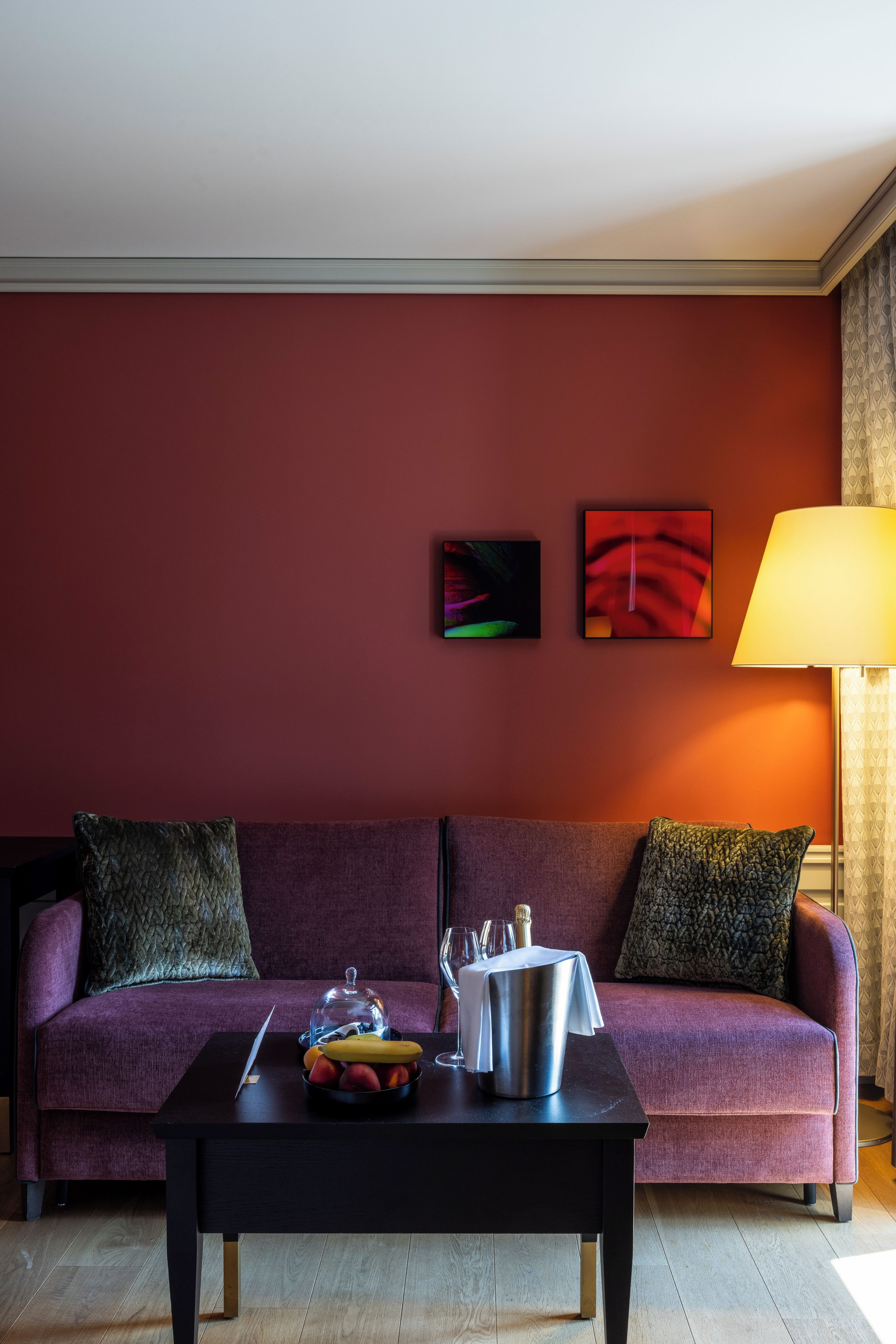 A room with a red wall, a purple sofa, a black coffee table with fruits and a champagne bottle, and two abstract paintings.