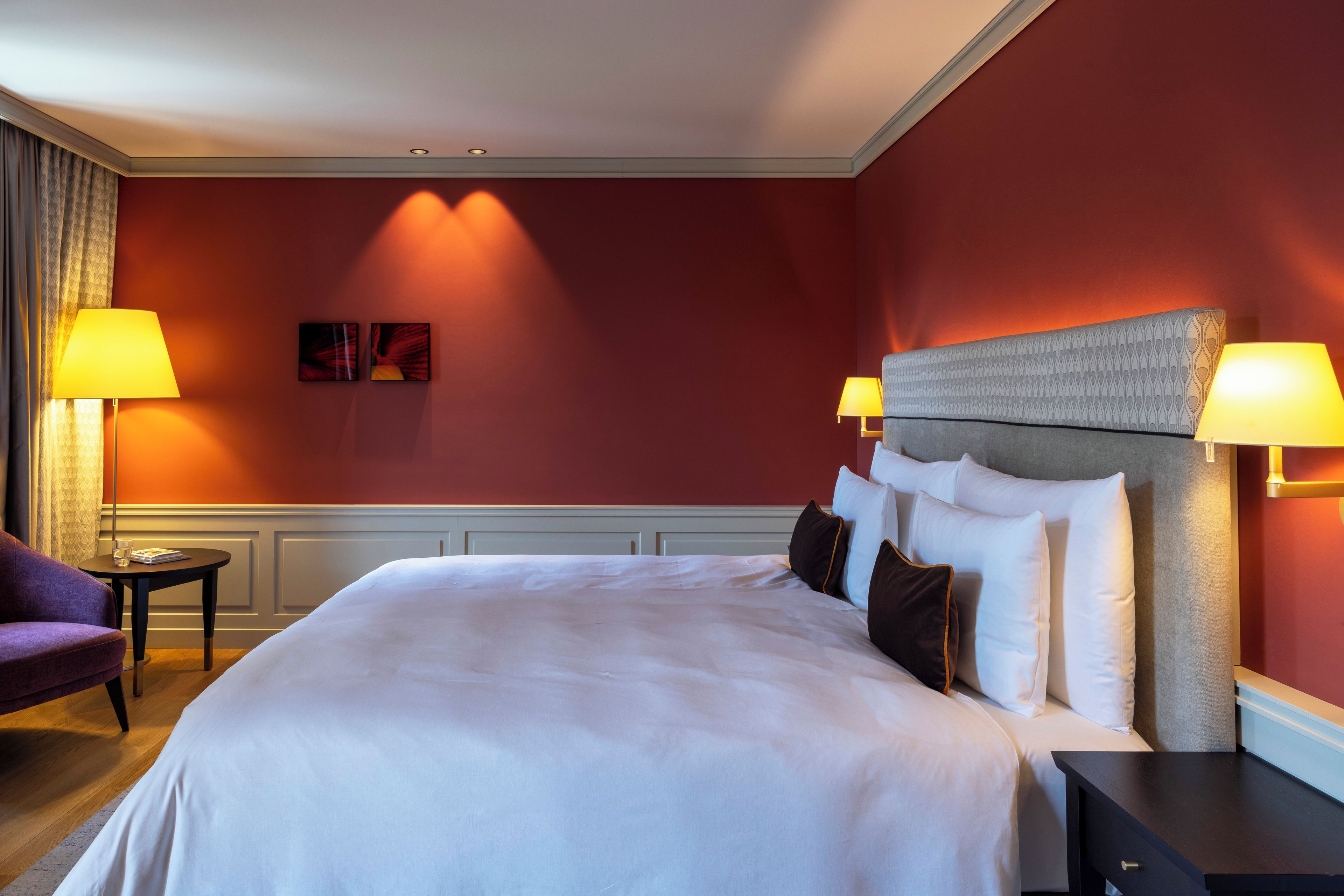 Habitación de hotel con una cama grande, dos lámparas de noche, una mesita pequeña y un sillón morado.