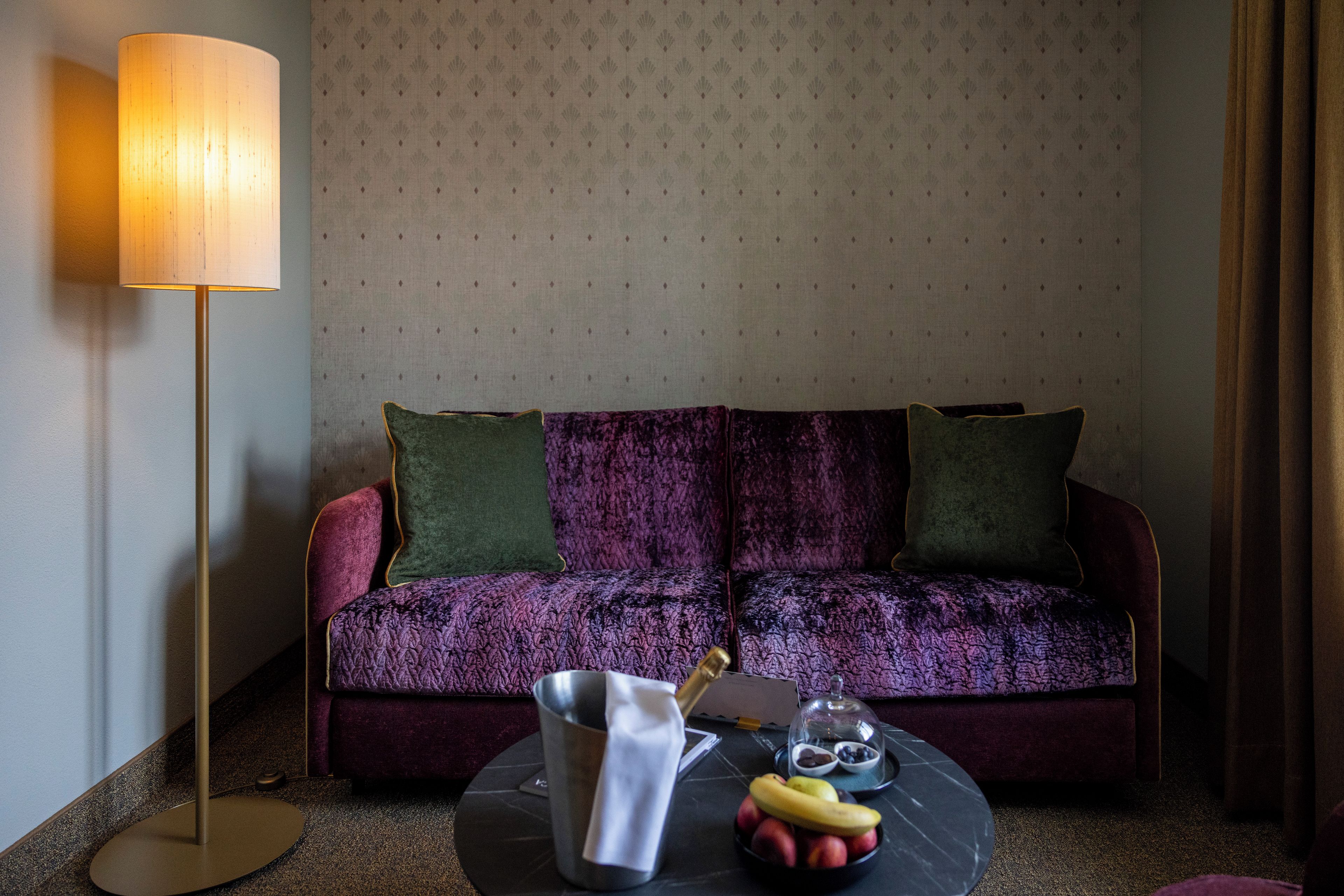 A room with a sofa, a lamp, a table with fruits and a cup, and patterned wallpaper.