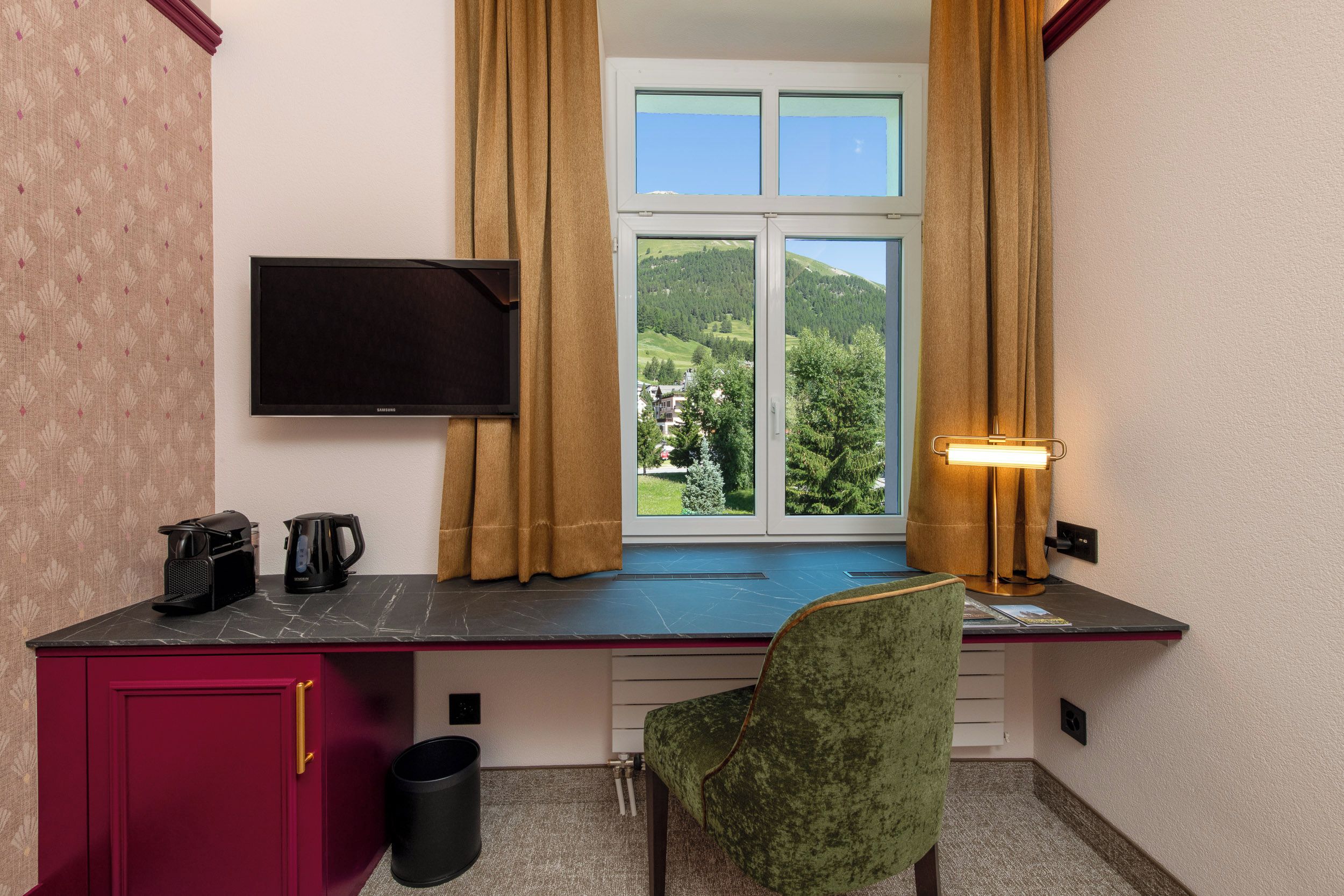 Ein Zimmer mit Schreibtisch, Stuhl, Fernseher, einem Fenster mit Vorhängen und einer Lampe.