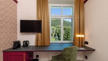Ein Zimmer mit Schreibtisch, Stuhl, Fernseher, einem Fenster mit Vorhängen und einer Lampe.