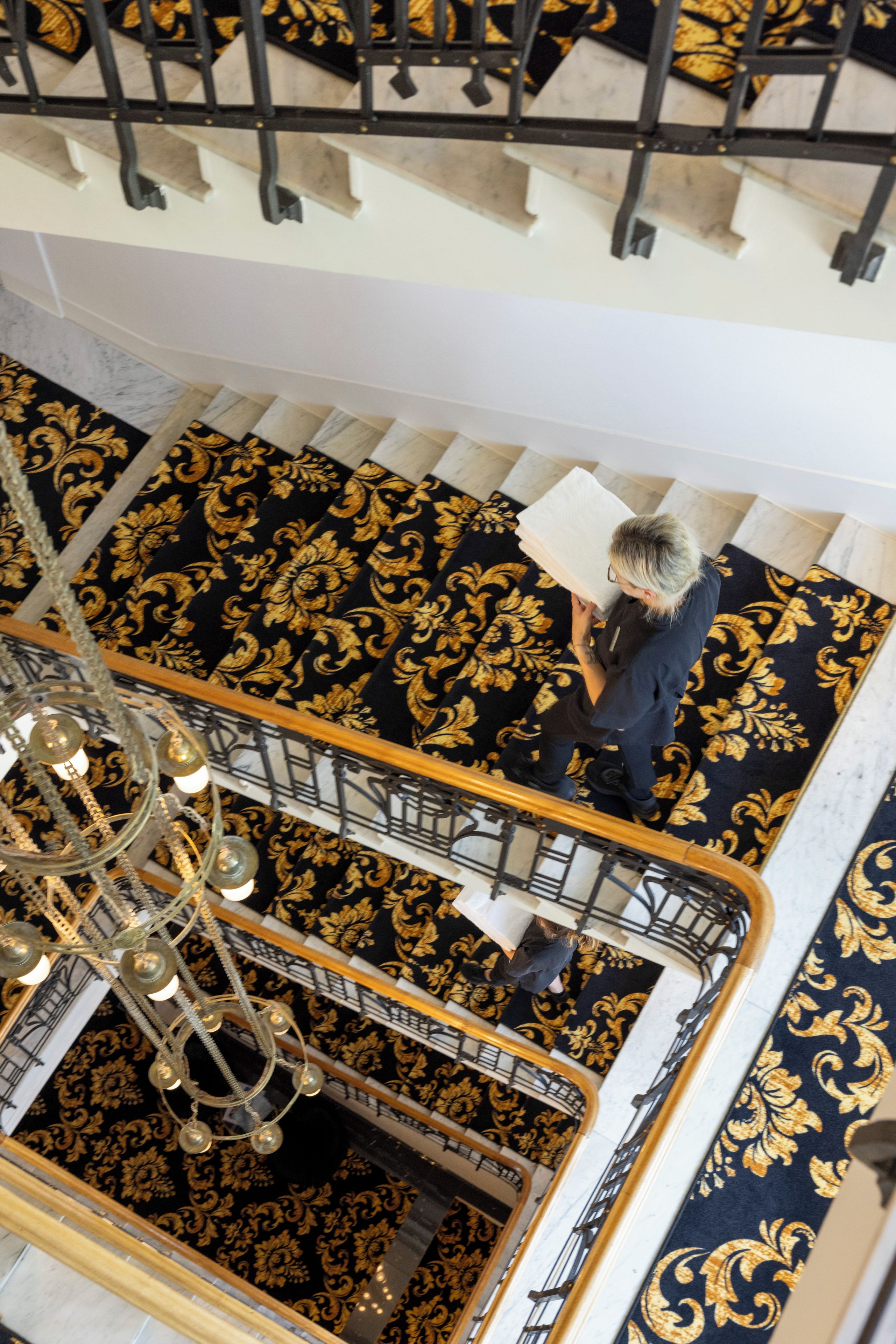 Eine Person steht auf einer Treppe mit aufwendigem, schwarz und goldfarbenem Teppich, einem Chandelier und einem weißen Geländer.