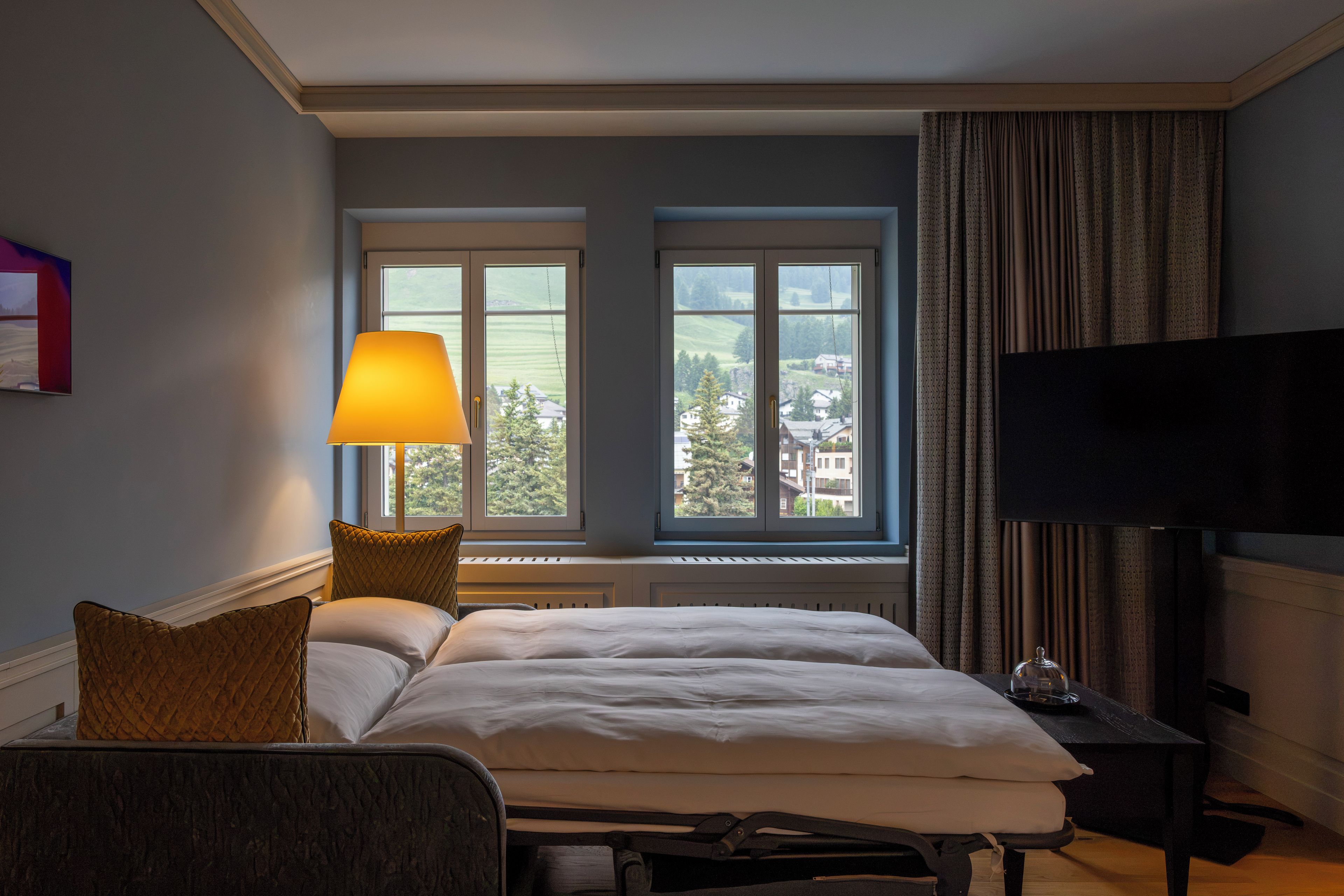 Une chambre d’hôtel avec un lit, une lampe de chevet, une télévision et une fenêtre donnant sur un paysage urbain.